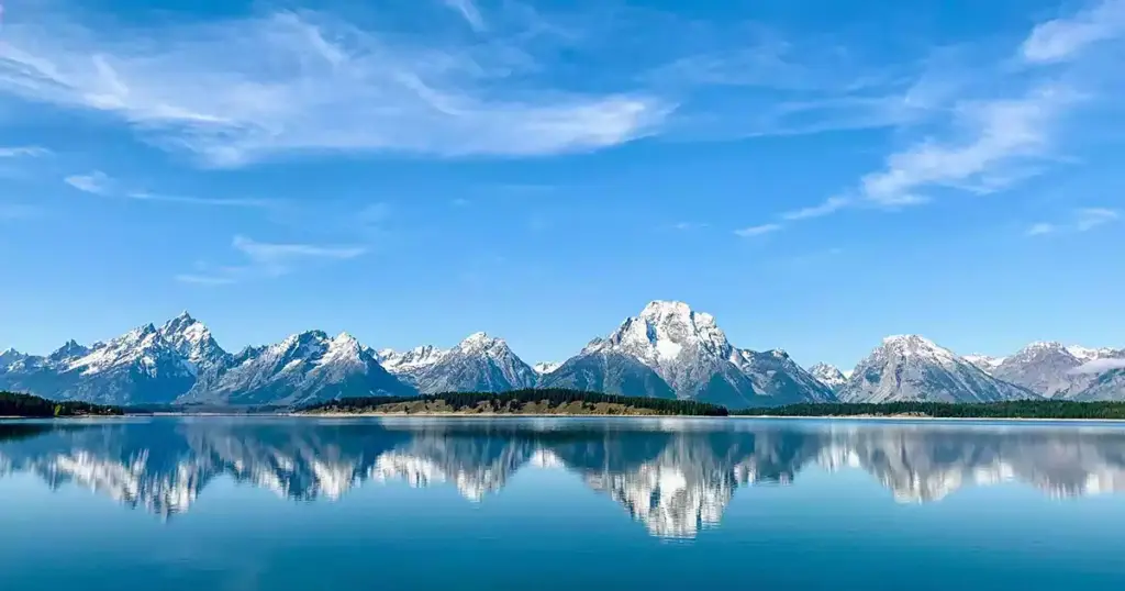 Teton Mountains reflecting in calm waters under a clear blue sky, showcasing the natural beauty of Jackson Hole.