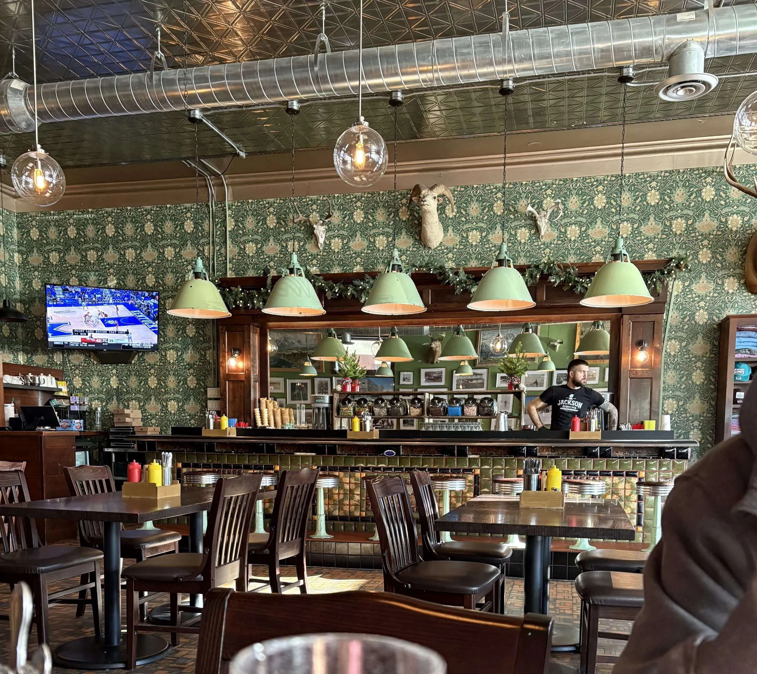 Interior of a restaurant featuring green pendant lights, a bar with condiments, a television displaying a sports game, and a staff member wearing a "Jackson" shirt, set against a patterned wallpaper and rustic decor, reflecting the dining atmosphere in Jackson Hole.