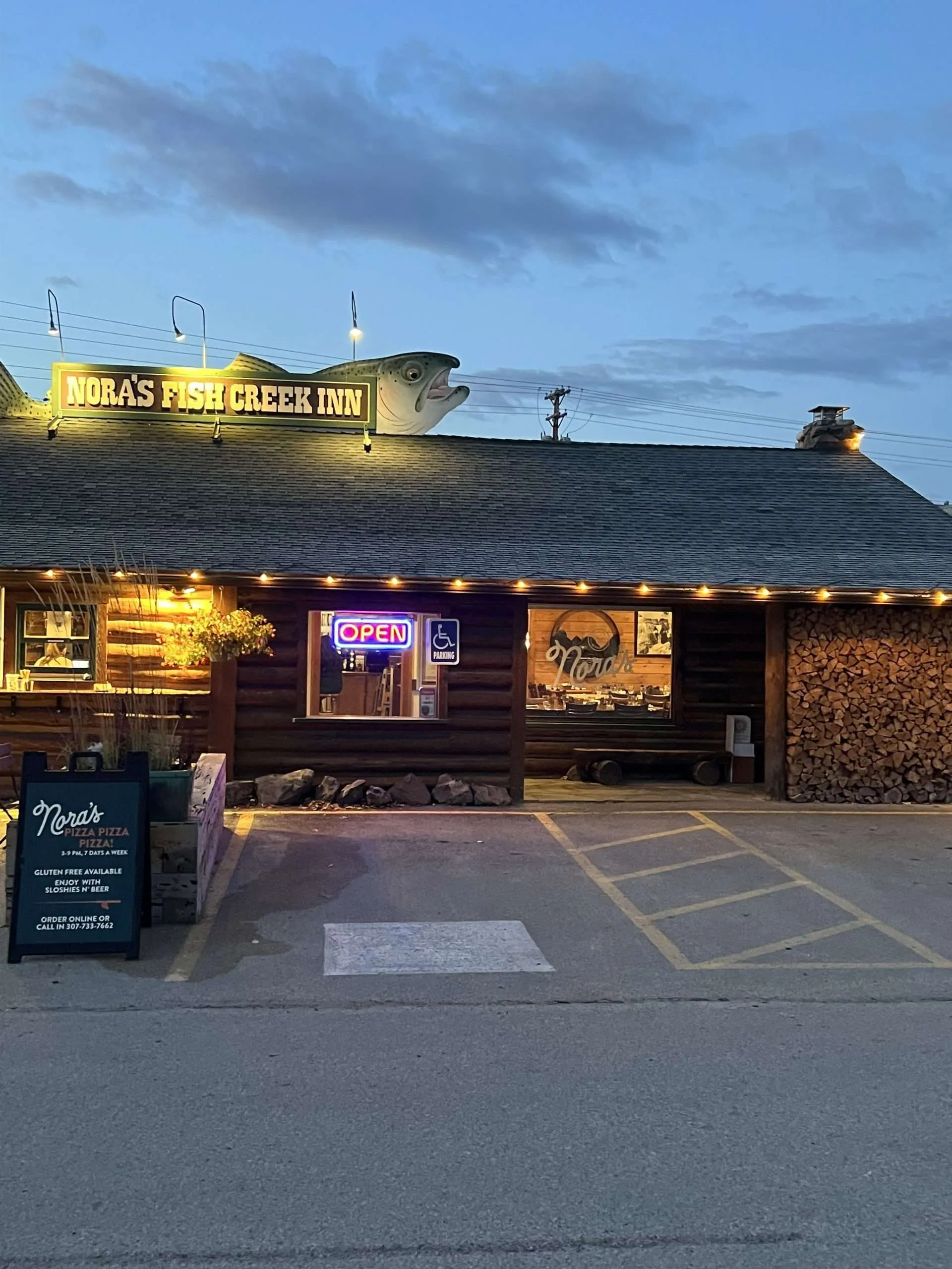 Nora's Fish Creek Inn sign featuring a large fish sculpture, rustic log cabin exterior, illuminated "OPEN" sign, and outdoor dining area in Jackson Hole.