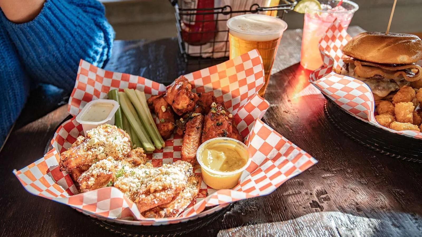 Basket of pub food featuring wings, celery sticks, dipping sauces, and a burger with tater tots, alongside craft beer and cocktails, in a family-friendly sports bar setting.