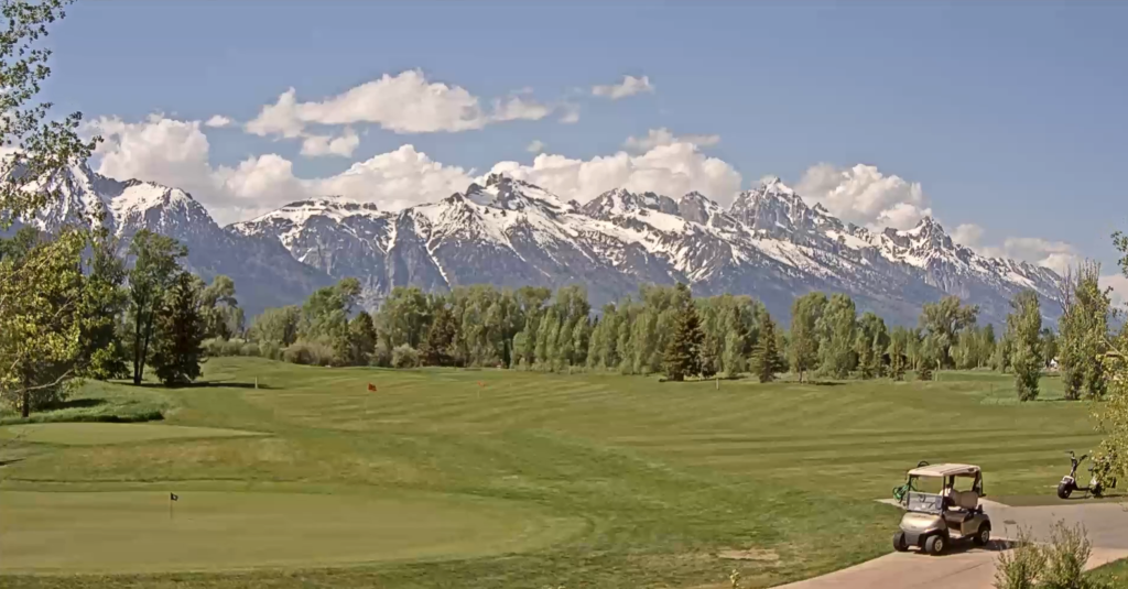 Golf course with manicured green and golf carts, overlooking the snow-capped Teton mountains under a blue sky with scattered clouds, representing outdoor activities in Jackson Hole.